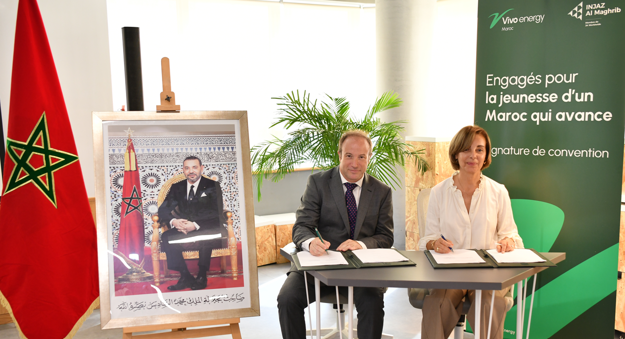 A man and woman sit next to each other behind a table, posed to sign an agreement. 