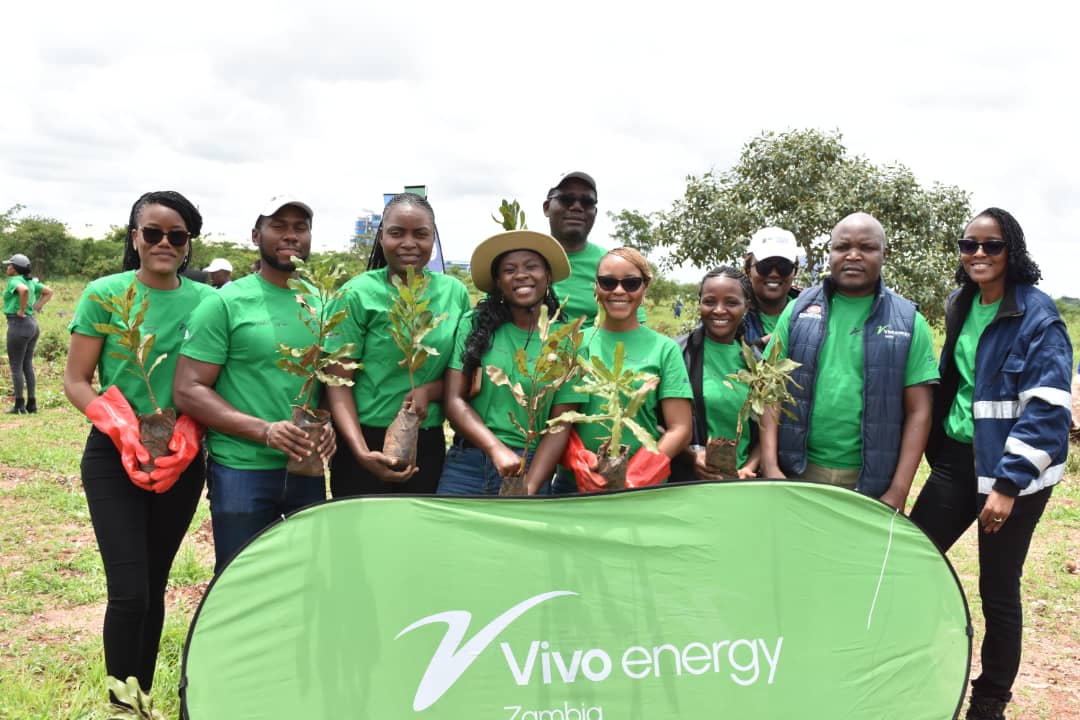 A group of 10 volunteers stand in a field behind a green banner, holding small saplings 