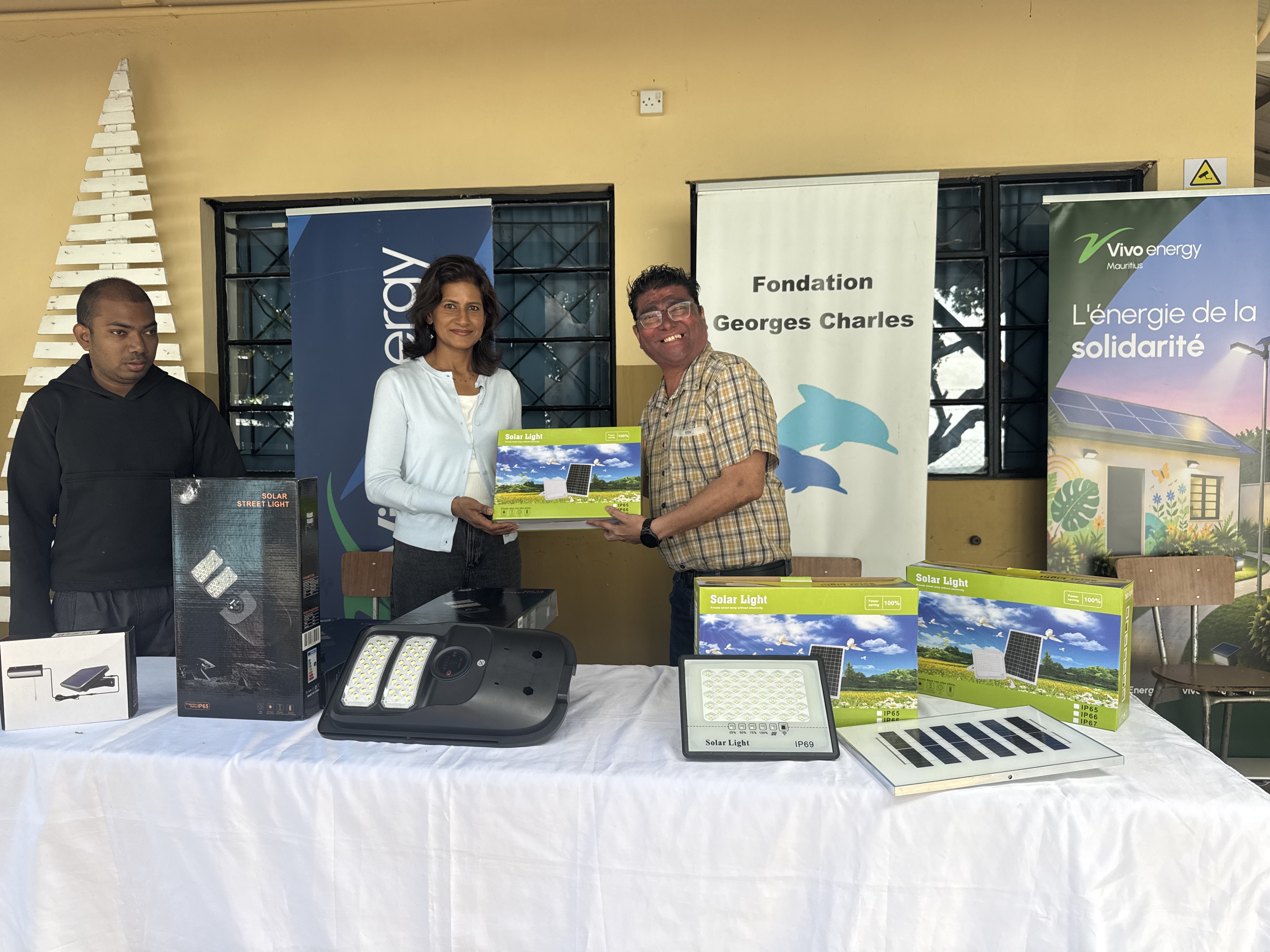 Derrière une table recouverte d’une nappe blanche, une collaboratrice de Vivo Energy Maurice et un représentant de la Fondation Georges Charles posent pour la caméra en tenant une lampe solaire emballée dans sa boîte.